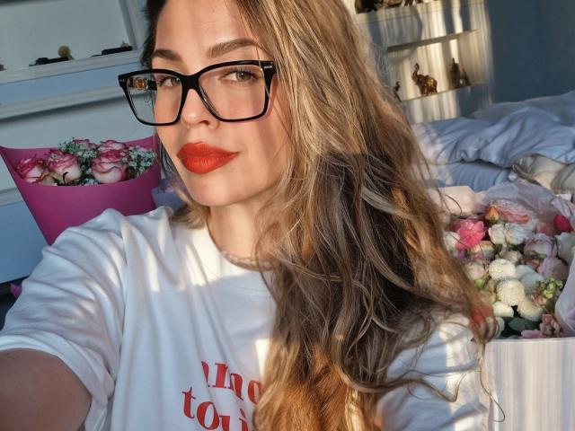 ValeriMair with long wavy blonde hair, black glasses, red lipstick, white shirt, looking at camera, holding pink and white rose bouquet, sunlight casting shadows.