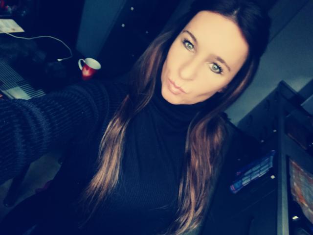 Una ragazza di nome Jordangirl, con lunghi capelli castani, occhi verdi e un'espressione accigliata, indossa un maglione nero a collo alto e sta scattando un selfie in una stanza poco illuminata, mostrando leggermente il seno..