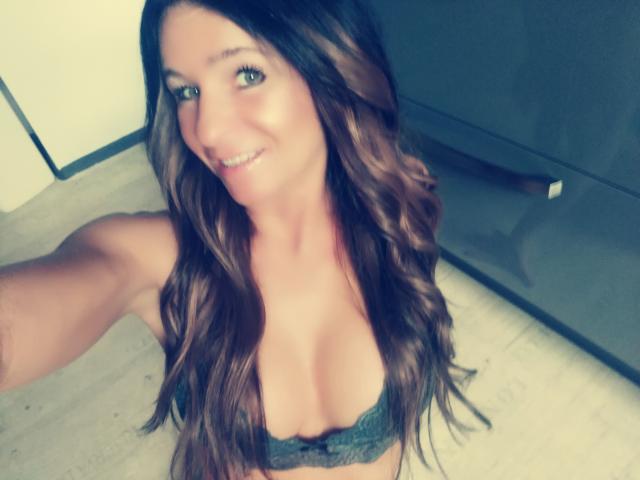 Jordangirl-atraente, com longos cabelos castanhos ondulados, olhos verdes, sorrindo, vestindo um sutiã de renda cinza, mostrando seus seios, tirando uma selfie em um quarto com pouca luz..