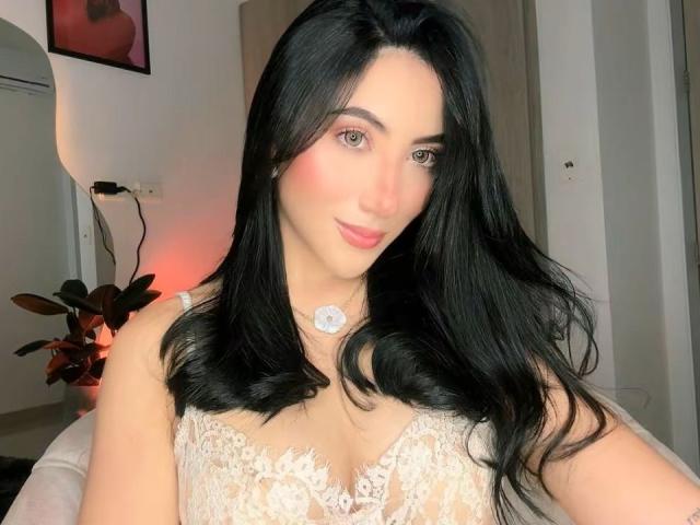 JuanaFox has long black hair, pink eyeshadow, and a slight smile. She's wearing a white lace dress with a floral necklace, showing her boobs. Her skin is fair.