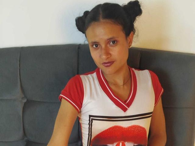 CrisyMarie with dark hair in double buns, wearing a red and white V-neck shirt with a lipstick print, sitting on a gray sofa, looking slightly to the right.