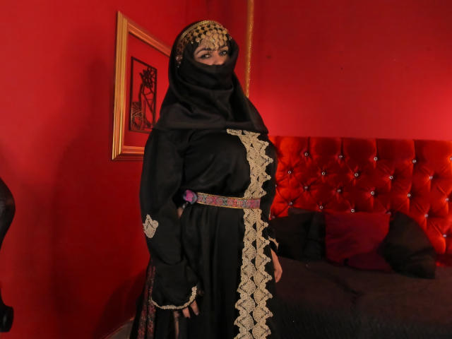 AnissaAli with black burqa, gold headpiece, and purple belt, standing in red room, showing her tits, next to red tufted bed with black and red pillows.