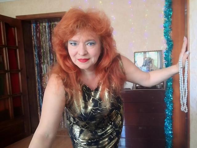 LadaMay with red, curly wig, blue eyeshadow, and gold patterned dress, holding pearl necklace, smiling, fair skin, small tits, standing in wooden room with blue tinsel.
