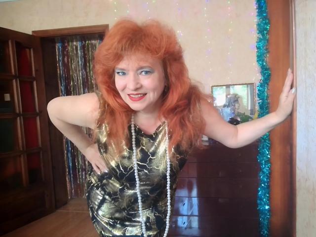 LadaMay with red curly hair, wearing a gold camouflage dress and long white necklace, smiling, leaning against a wall, hands on hips, blue tinsel on the right.