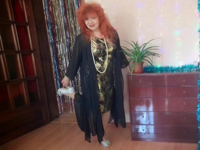 LadaMay with red hair, black sheer robe, gold dress, long necklace, white heels, holding white purse, standing next to wooden dresser with green plant, smiling.