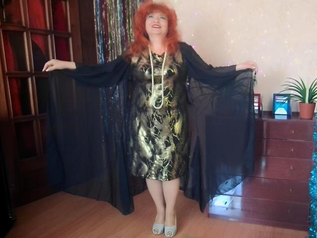 LadaMay with red hair, black sheer robe, gold patterned dress, necklace, and light blue shoes, standing with arms out, smiling, wooden floor, small plant on dresser.