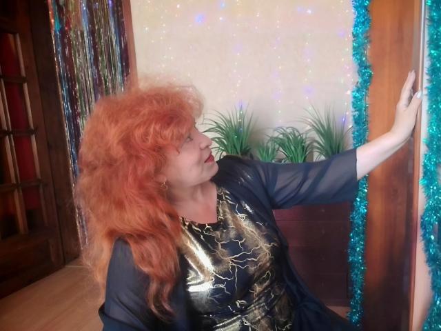 LadaMay with curly red hair, black sheer top, and shiny gold patterned shirt, leaning on a door with blue tinsel, her hand raised, looking to the side.
