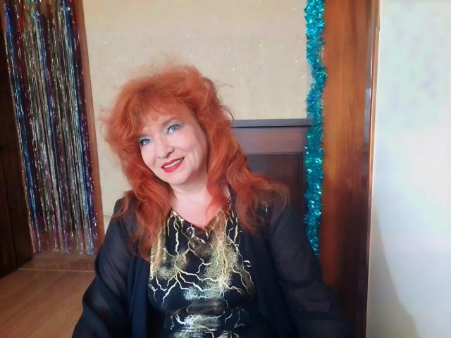 LadaMay with red curly hair, blue eyes, and red lipstick, wearing a black sheer top with gold lightning pattern, and blue tinsel, smiling at the camera.