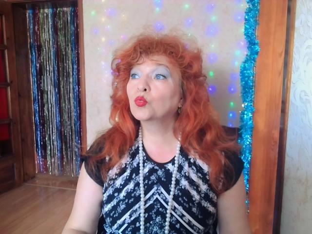 LadaMay with curly red hair, blue eyeshadow, and red lipstick, pouting her plump lips, wearing a black and white patterned top, sitting in front of colorful lights.
