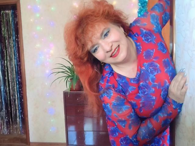 LadaMay with curly red hair, blue eyes, red lipstick, wearing a red floral dress, leaning forward, smiling, showing her big tits, in a colorful room with sparkles.