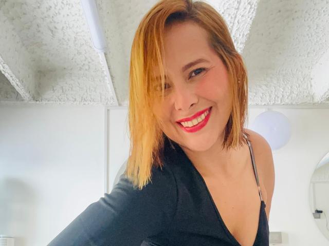 Sarita Velez mit kurzen, blonden bis braunen Ombre-Haaren, lächelnd mit leuchtend rotem Lippenstift, trägt ein schwarzes Oberteil, das ihre nackten Schultern und kleinen Brüste zeigt..