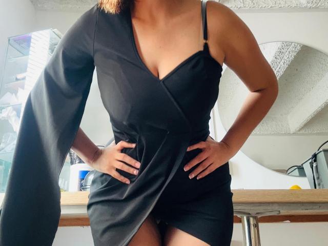 Sarita Velez in un abito nero con un profondo scollo a V, che mette in mostra il seno, con le mani sui fianchi, in piedi con le gambe leggermente divaricate, con le bretelle nere visibili..