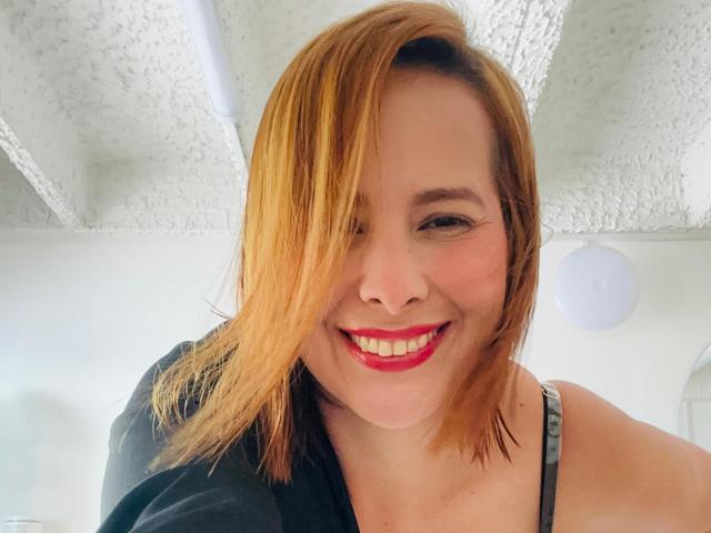 SaritaVelez with red lipstick, smiling, showing teeth, black top, brown shoulder-length hair, white textured ceiling, white ceiling light, smiling at camera, upper body shot.