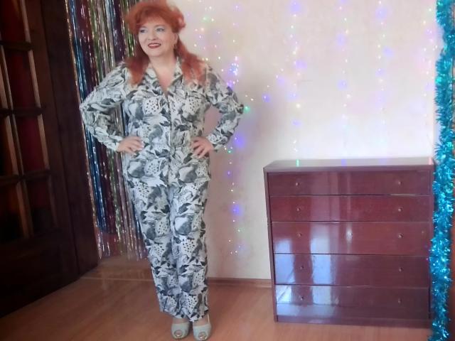 LadaMay with red hair, wearing a floral jumpsuit, standing with hands on hips, wearing white heels, in front of a wooden dresser, with colorful sparkles and tinsel.