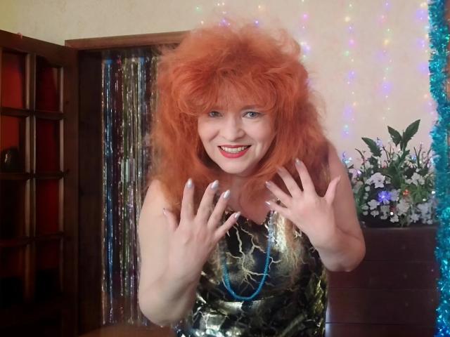 LadaMay with wild red hair, smiling, hands raised, wearing a shiny black dress, showing her tits, in a colorful, festive room.