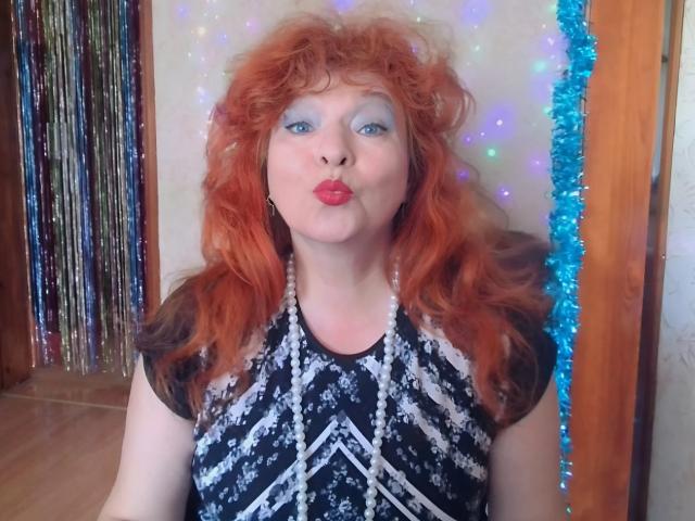 LadaMay with curly red hair, blue eye makeup, and red lipstick, pouting her lips, wearing a black and white patterned dress, standing in front of blue and purple fairy lights.