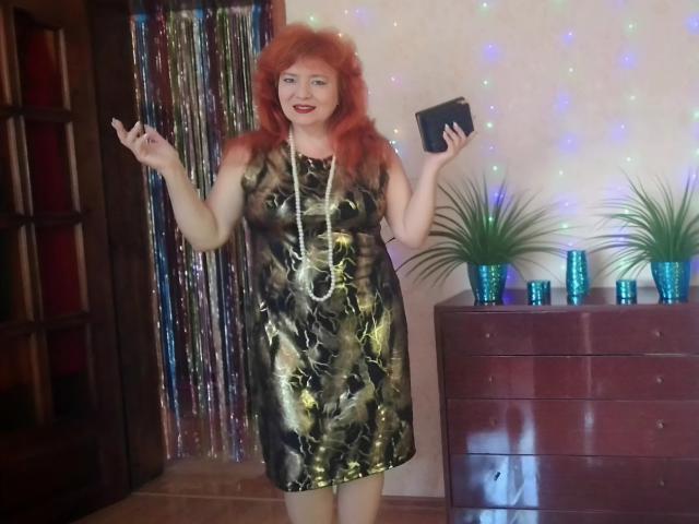 LadaMay with red hair, green and black patterned dress, necklace, holding phone, raised arms, standing by wooden dresser with green plants and blue candles, smiling.