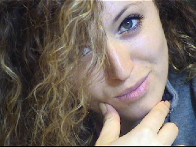 SassyKate with curly brown hair, blue eyes, and a slight smile, touching her chin with her right hand, showing a cute and relaxed expression.