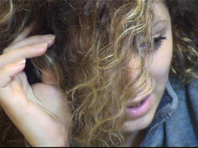 SassyKate has curly, blonde hair covering her face, with a hand touching her head. Her eyes are slightly closed, and she's wearing a gray shirt.