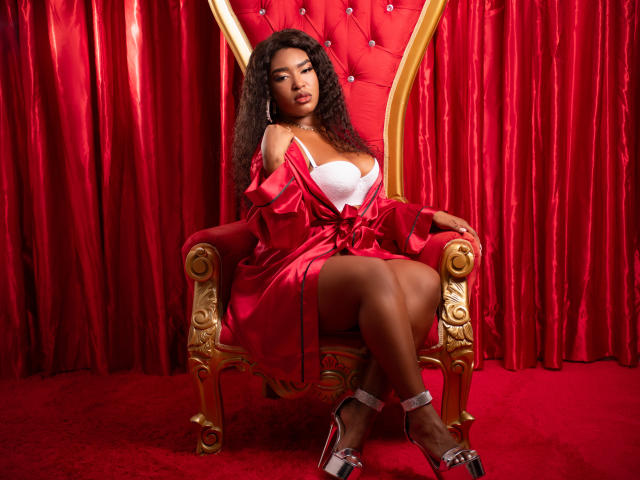 ZairaWarner with curly black hair, red satin robe, white bra, showing her tits, sitting on gold throne, red carpet, silver heels, red curtains, looking at camera.