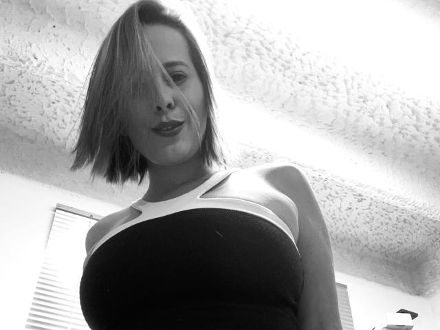 Black-and-white photo of SaritaVelez with a bob haircut, wearing a strapless top, her tits prominent, smiling slightly, ceiling with textured pattern above her head.