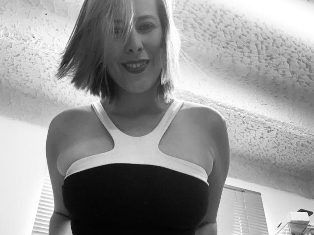 Fotografia in bianco e nero di Sarita Velez con un taglio di capelli corto, che sorride, indossa un top che mette in mostra il suo seno prosperoso, ed è in piedi in una stanza con un soffitto strutturato..