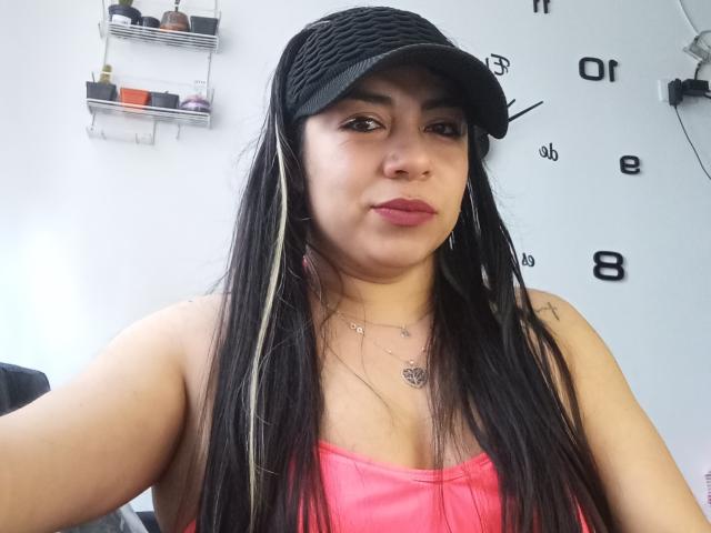 EvaLunaLatina with long black and blonde hair, wearing a black cap, pink tank top, and necklace, has small tits and a cute face, standing in a room.