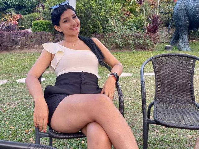 EmiliRiders69 with tan skin, black pigtails, white ruffled top, black high-waisted shorts, crossed legs, sitting on metal chair, smiling, sunglasses on head, green garden background.