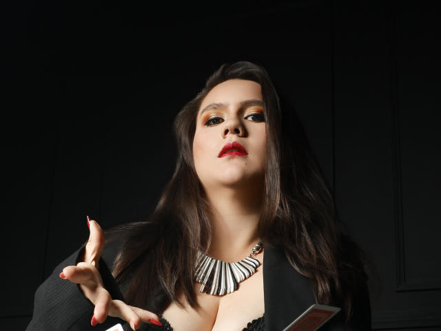 AvienLume with dark brown hair, red lipstick, black dress, zebra necklace, and red nails, pointing with right hand, showing large, prominent tits. Dark background.