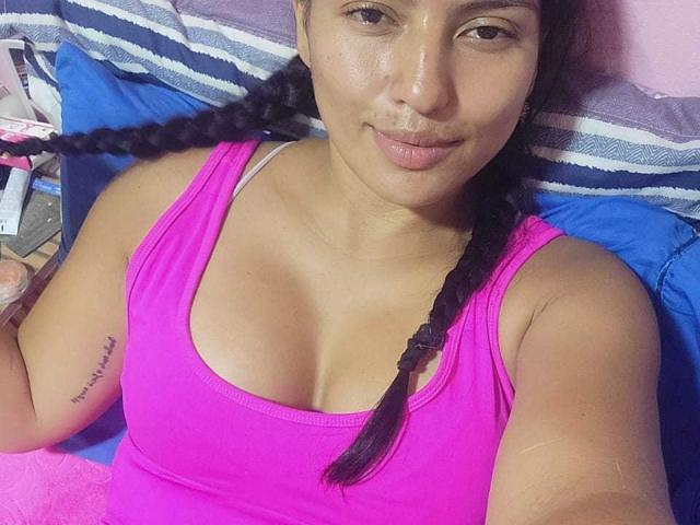 TamaraOriz has brown skin, small tits, and a cute face with braided black hair, wearing a pink tank top, lying on a bed, looking at the camera.