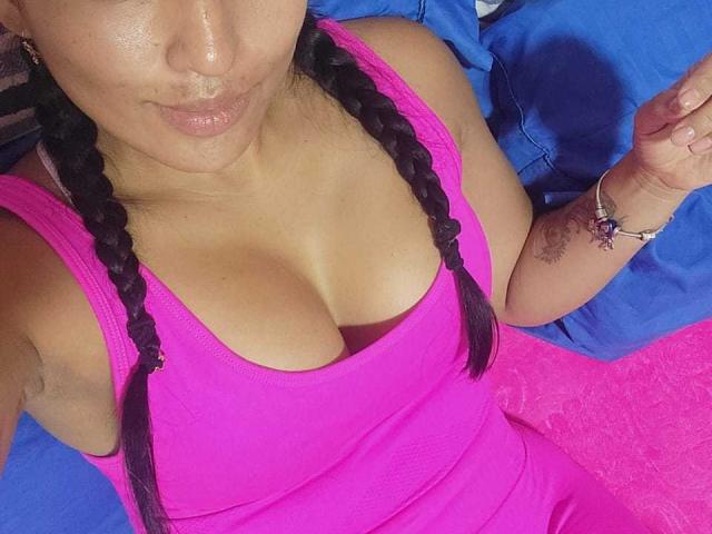 TamaraOriz with braided black hair, pink tank top showing her big tits, blue and pink background, subtle tattoo on her right wrist, and a pink towel.