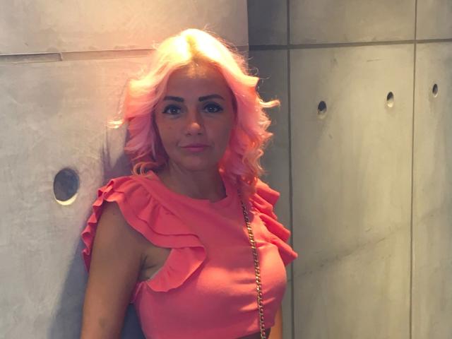 CoconutCandy-hot with pink wavy hair, wearing a ruffled pink top, standing against a gray wall. She's got a cute face, small boobs, and a gold chain.