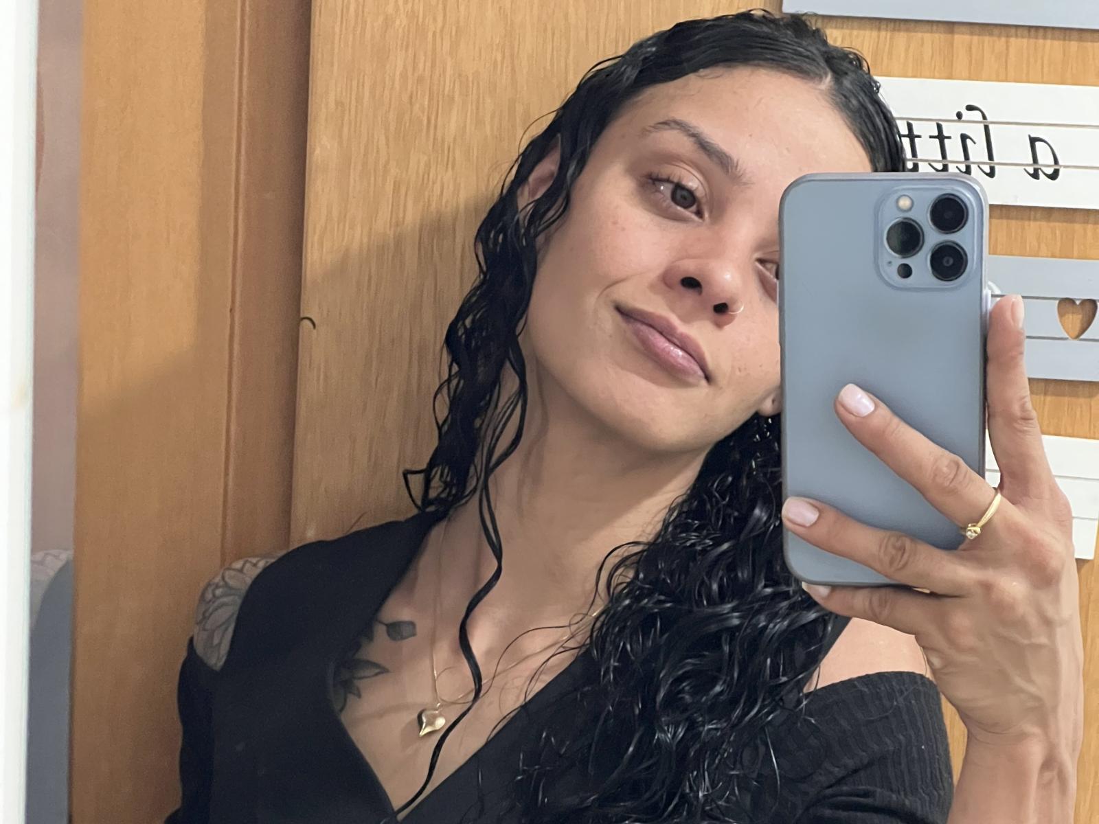 Gatinha69 com cabelo preto molhado, pequenas tatuagens no ombro direito, segurando um iPhone azul, vestindo uma camiseta preta com decote em V, sorrindo levemente, mostrando um pequeno anel de ouro no dedo..