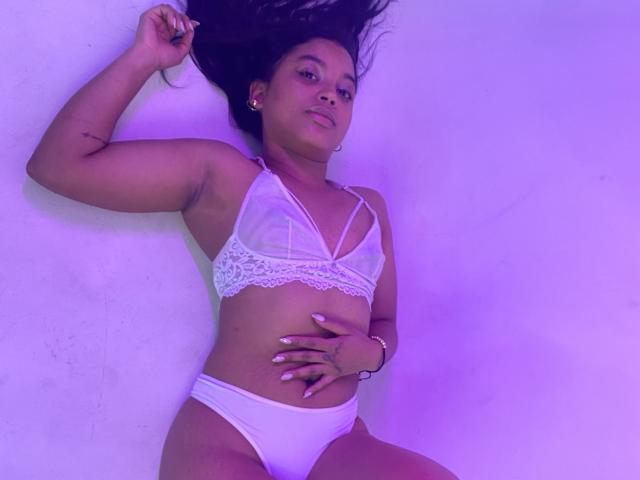 HilaryCook lying down, wearing white lace bra and panties, hand on her belly, purple light, dark hair, small tattoo on arm, confident expression, slight smile.