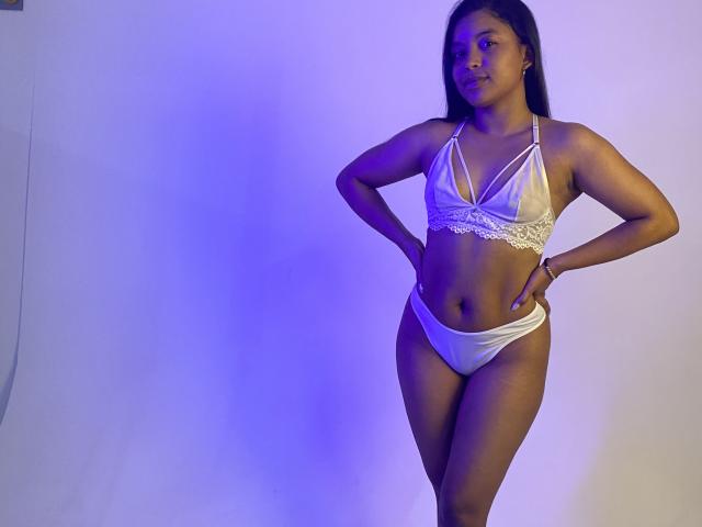 HilaryCook stands with hands on hips, wearing a white lace bra and matching panties. She has a medium skin tone, black hair, and a confident, sexy look.