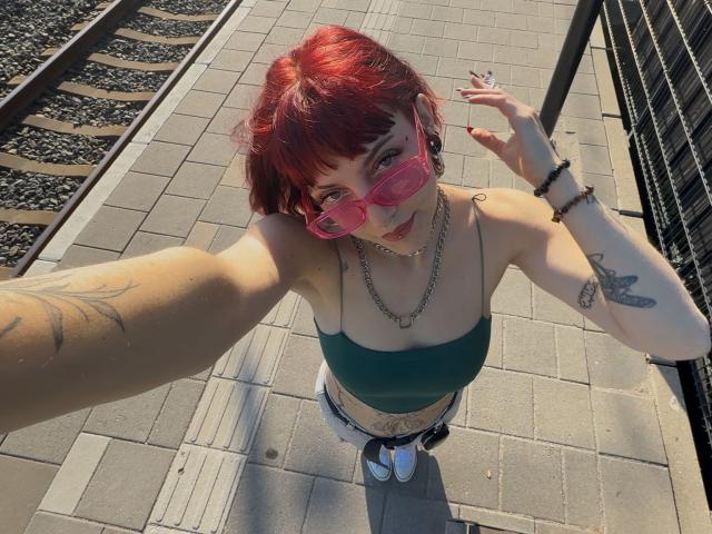 MistiqueLeyla with red hair, pink sunglasses, green top, tattoos, chain necklace, and white shorts, taking a selfie from above, showing her small tits and white sneakers.
