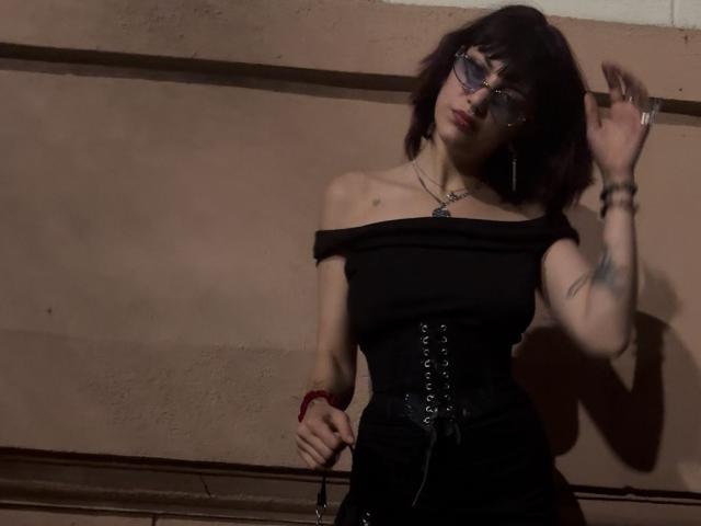 MistiqueLeyla with dark bob, glasses, black off-shoulder top, corset, black skirt, tattoos, red bracelet, standing, hand up, shadowed, looking slightly up, dark brown wall background.