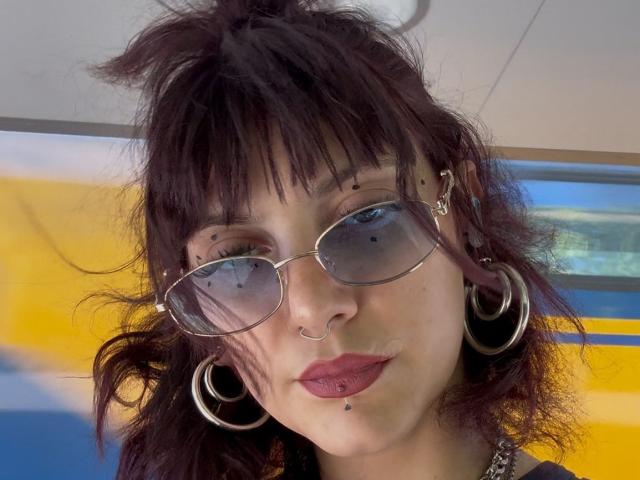 MistiqueLeyla with messy dark hair, bangs, nose ring, multiple facial piercings, silver aviator sunglasses, hoop earrings, and pink lipstick, looking serious.