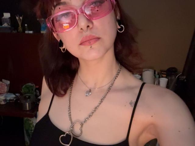 MistiqueLeyla with pink glasses, brown hair, nose ring, lip ring, black top, heart necklace, and earrings. Her tits are small, her skin is pale, and she's looking serious.
