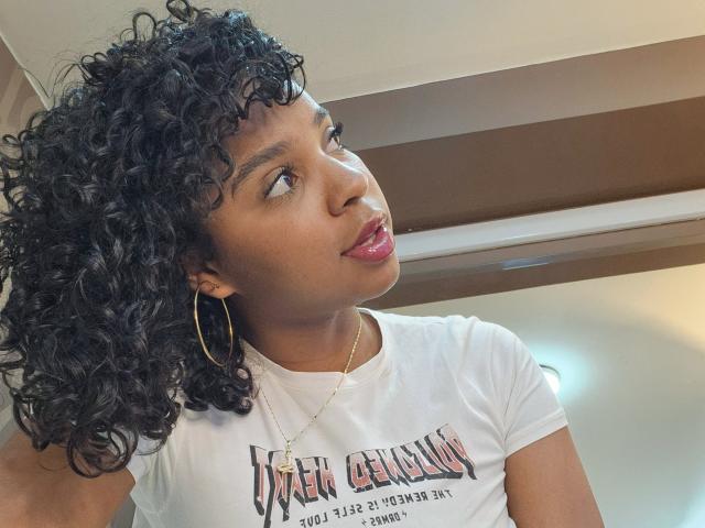 LisaMendez with curly black hair, wearing a white tee, gold hoop earrings, and pink lipstick, looking up with a slightly open mouth. She's got a cute brown face.