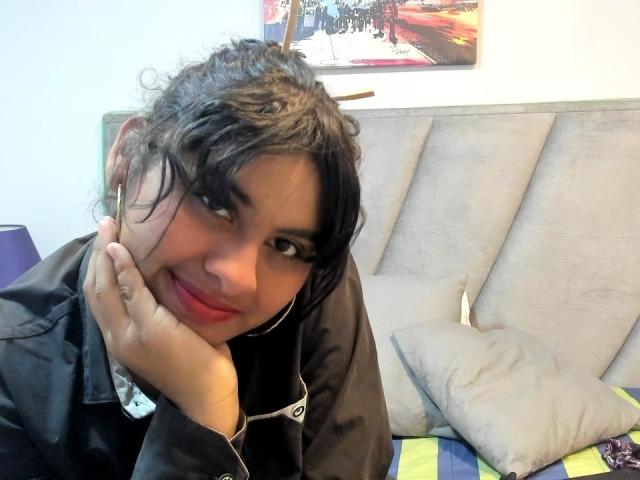AnanyaSyed with curly black hair, red lipstick, and black jacket, smiling at the camera, hand under chin, white earbuds, on a gray couch with a green-striped pillow.