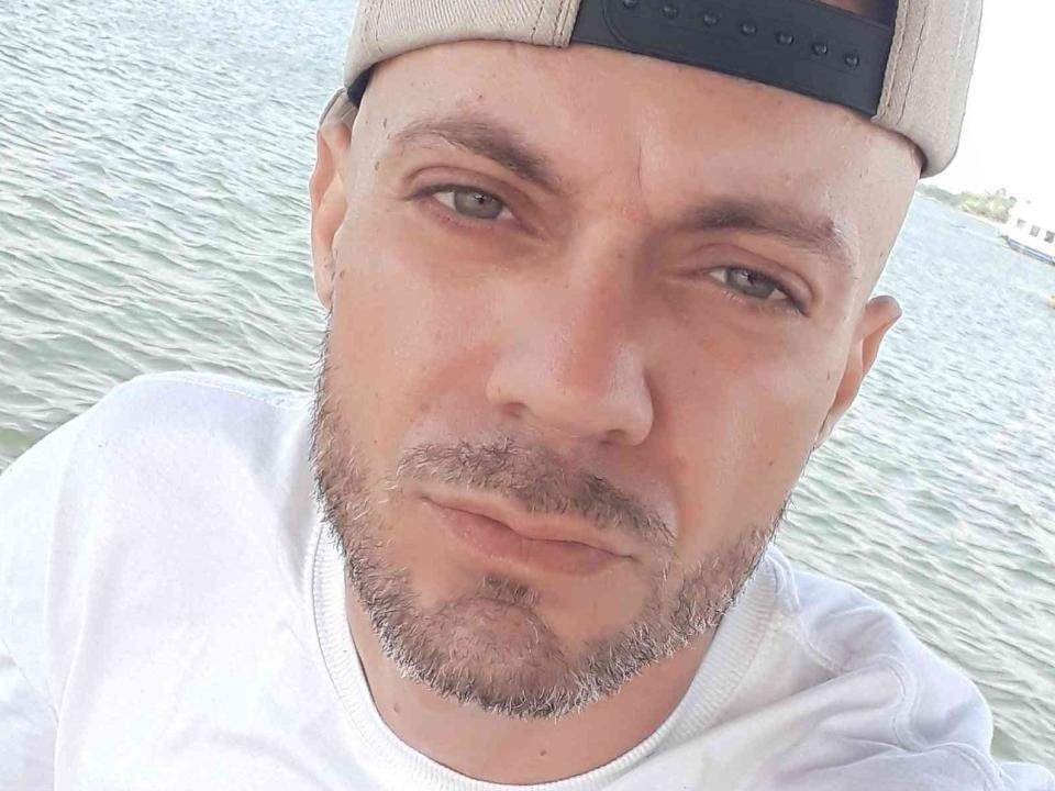 Cadu69Centim, com uma expressão séria, olhos castanho-claros, barba curta, camiseta branca e boné de beisebol bege, está em pé perto de uma massa de água azul-esverdeada. Seu nariz é ligeiramente arrebitado..