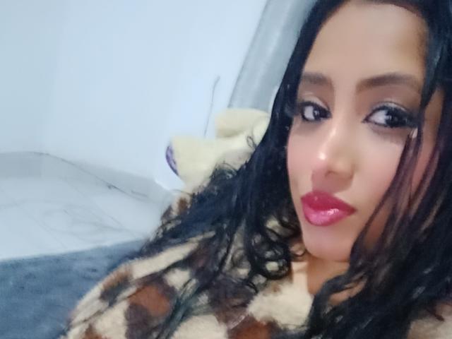 MariaCatalina has dark curly hair, pink lipstick, and is wearing a brown and beige fur coat. Her makeup is smoky, and she's looking at the camera with a slight smile.