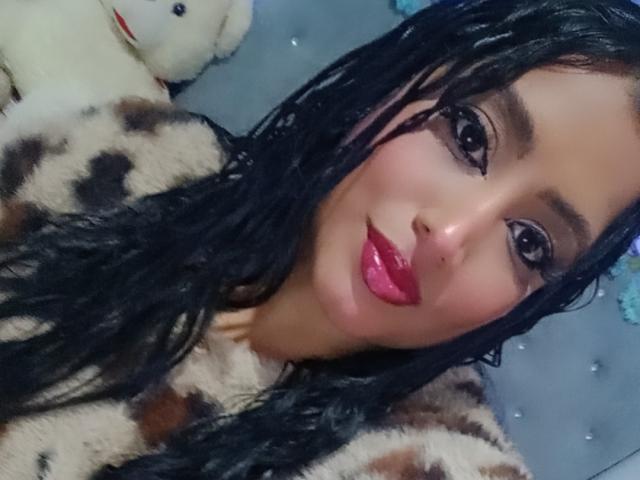 MariaCatalina with wet black hair, red lipstick, and a fur coat, showing off her pouty ass-adjacent face in a selfie, with a stuffed toy in the background.