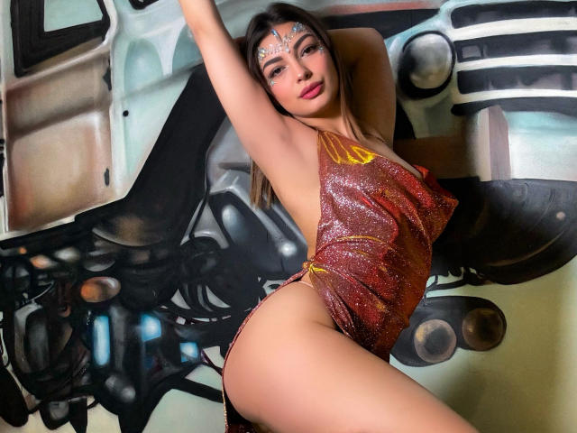 AmbreLaNebulleuse with sparkly red dress, exposing her ass, arms raised, wearing a jeweled forehead piece, brown hair, and red lipstick, in front of a graffiti wall.