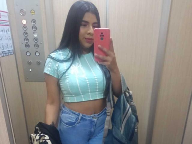 CharlotteXStone, con lunghi capelli neri, indossa una canottiera a righe azzurra e jeans blu, tiene in mano un telefono rosa e si mostra con il seno e i glutei in un selfie allo specchio di un ascensore..