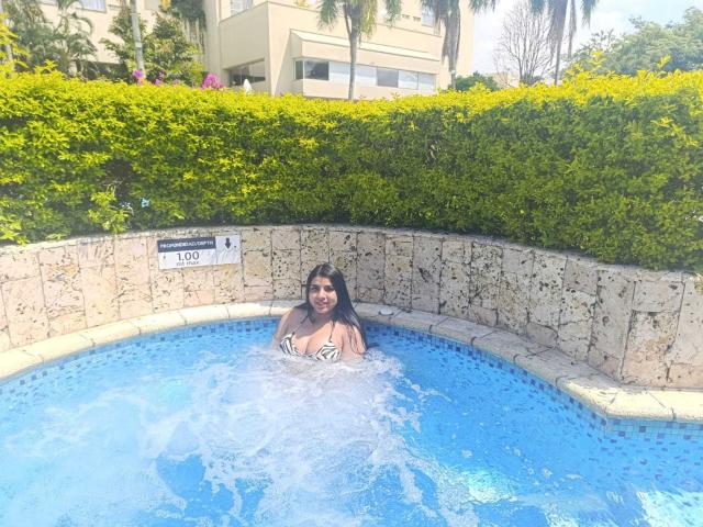 CharlotteXStone with large boobs, dark hair, and a smile, sitting in a small blue pool, water splashing around her, wearing a white and black bikini top.