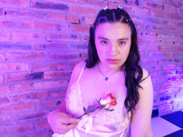 TaraTaylor with dark hair in pigtails, wearing a white lace camisole, holding a rose over her tits, blue necklace, pink and purple lighting, brick wall in background.
