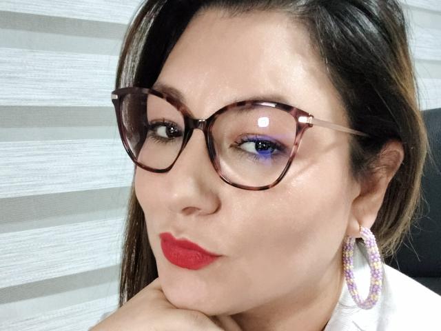 Sakoto Dika a les cheveux brun foncé, porte des lunettes à monture écaille de tortue, du maquillage pour les yeux violet, du rouge à lèvres rouge, et des boucles d'oreilles blanches avec des perles violettes, et repose son menton sur sa main..