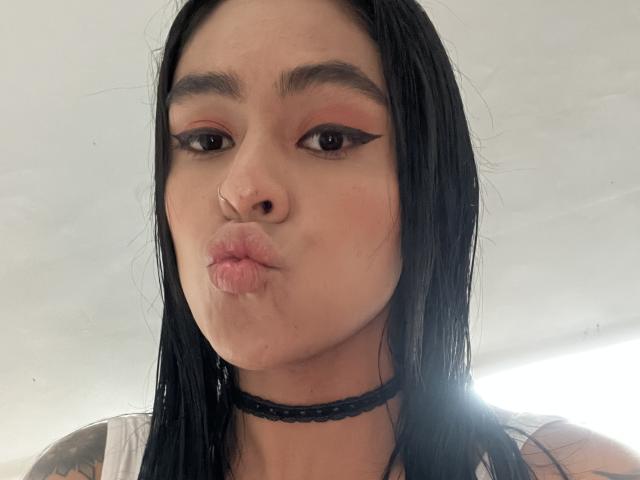 StellaBeckker with black hair, wearing a choker, puffy pink lips, and winged eyeliner, making a puckering face. She has a small nose ring and tattoo on her arm.
